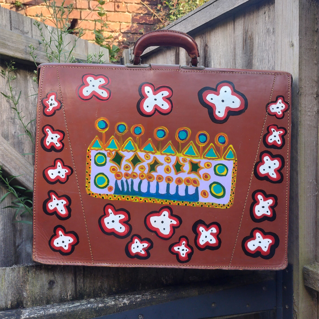 Back of It's shit now the Queen has gone piece on vintage briefcase. Flowers around the edges white, edged in red and then black with black dots on them. In the middle is hand painted image of a crown.