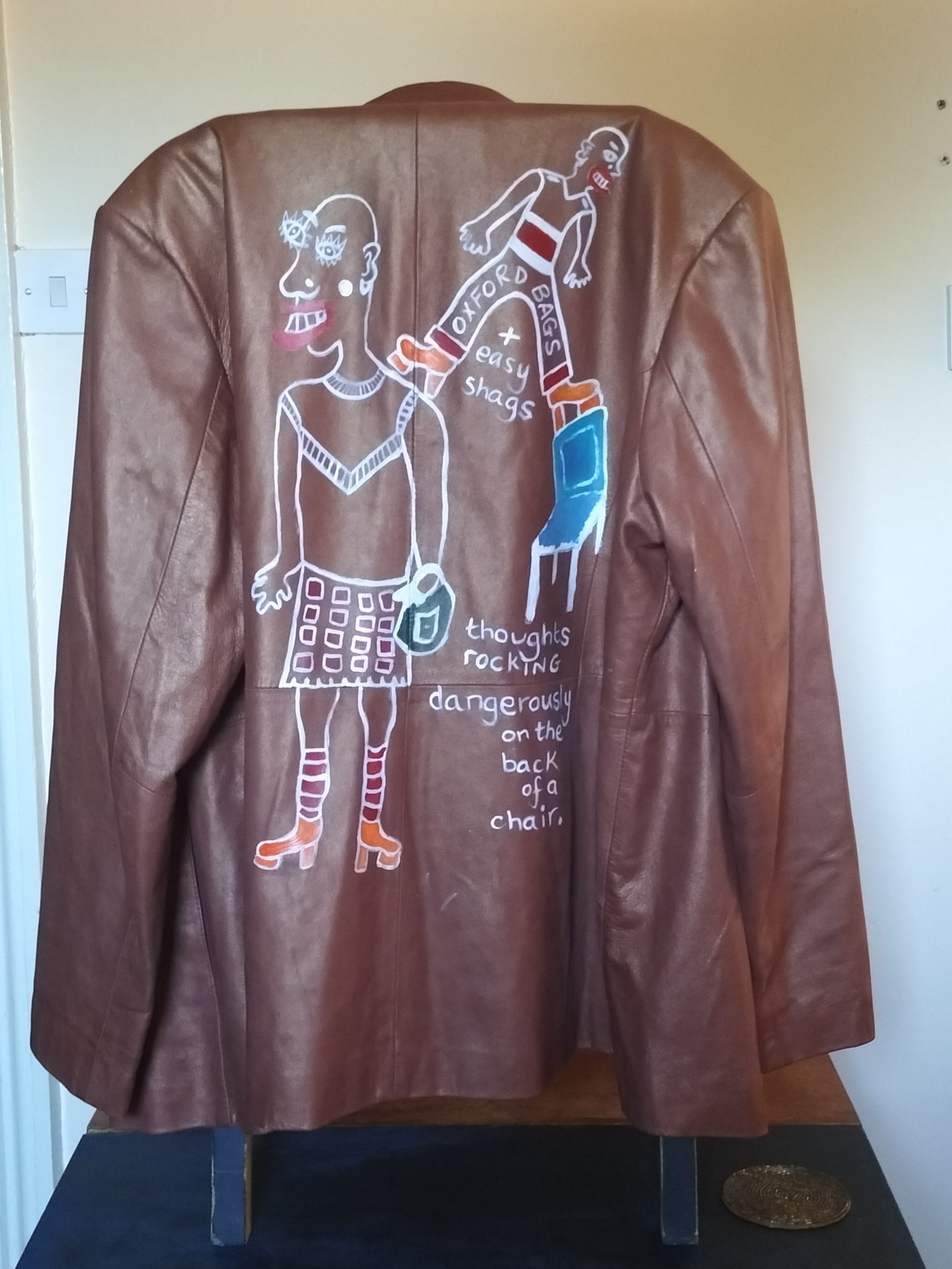 Size 16 hand painted leather Arma brown jacket. Hand painted on the back of the jacket. Person on the left of the image wears a skirt and platform orange shoes with stripey knee length socks and is carrying a bag. Smaller figure stands on the back of a chair that appears to be rocking and one foot is on the shoulder of the larger figure and the other on the back of the chair. The smaller figure has the words Oxford Bags across their trousers and below that is the letter + easy shags. Words further down the image read "thoughts rocking dangrously on teh back of a chair". Lovely soft leather. Buttoned front of jacket. One off original.