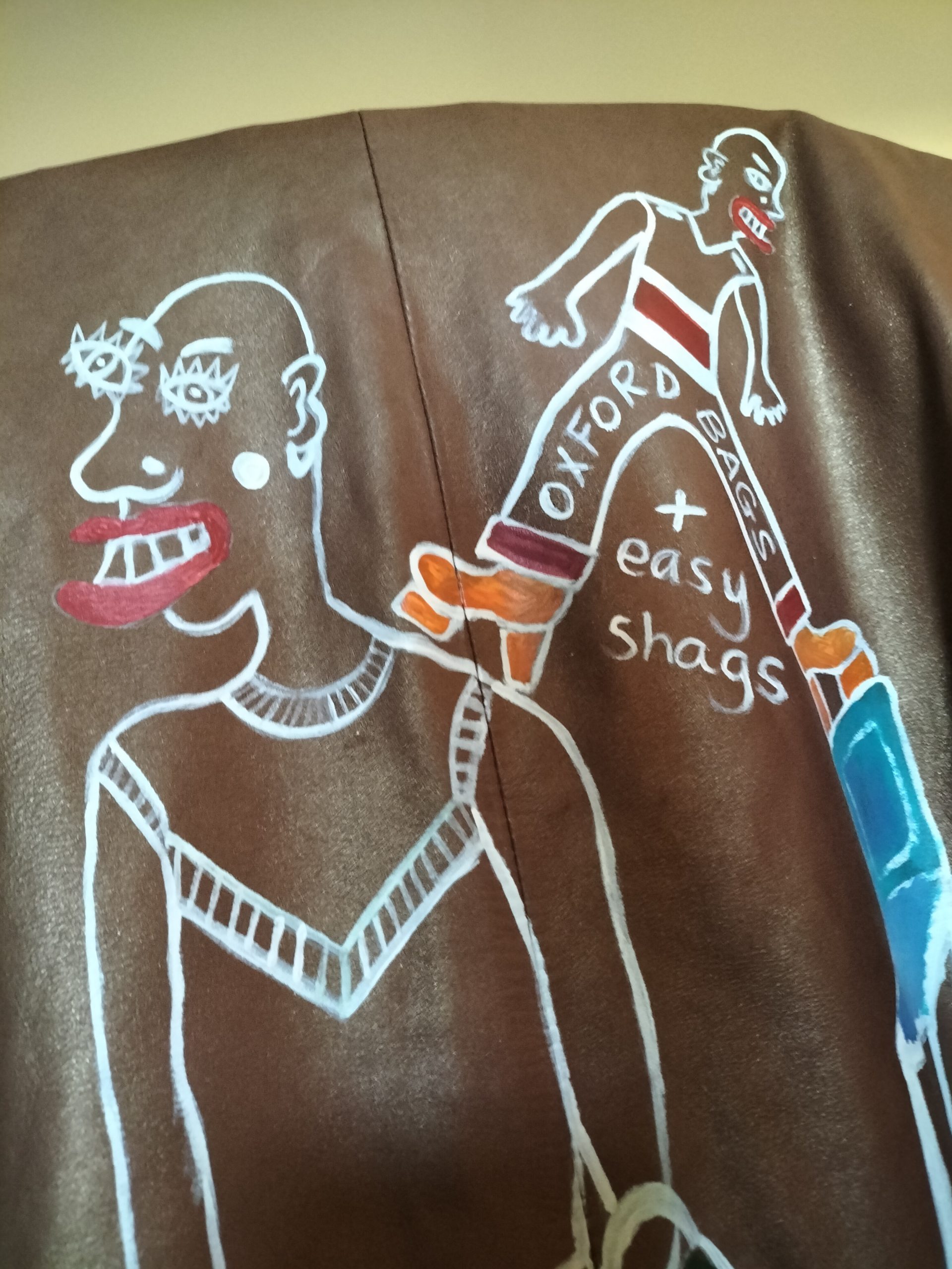 Size 16 hand painted leather Arma brown jacket. Hand painted on the back of the jacket. Person on the left of the image wears a skirt and platform orange shoes with stripey knee length socks and is carrying a bag. Smaller figure stands on the back of a chair that appears to be rocking and one foot is on the shoulder of the larger figure and the other on the back of the chair. The smaller figure has the words Oxford Bags across their trousers and below that is the letter + easy shags. Words further down the image read "thoughts rocking dangrously on teh back of a chair". Lovely soft leather. Buttoned front of jacket. One off original.