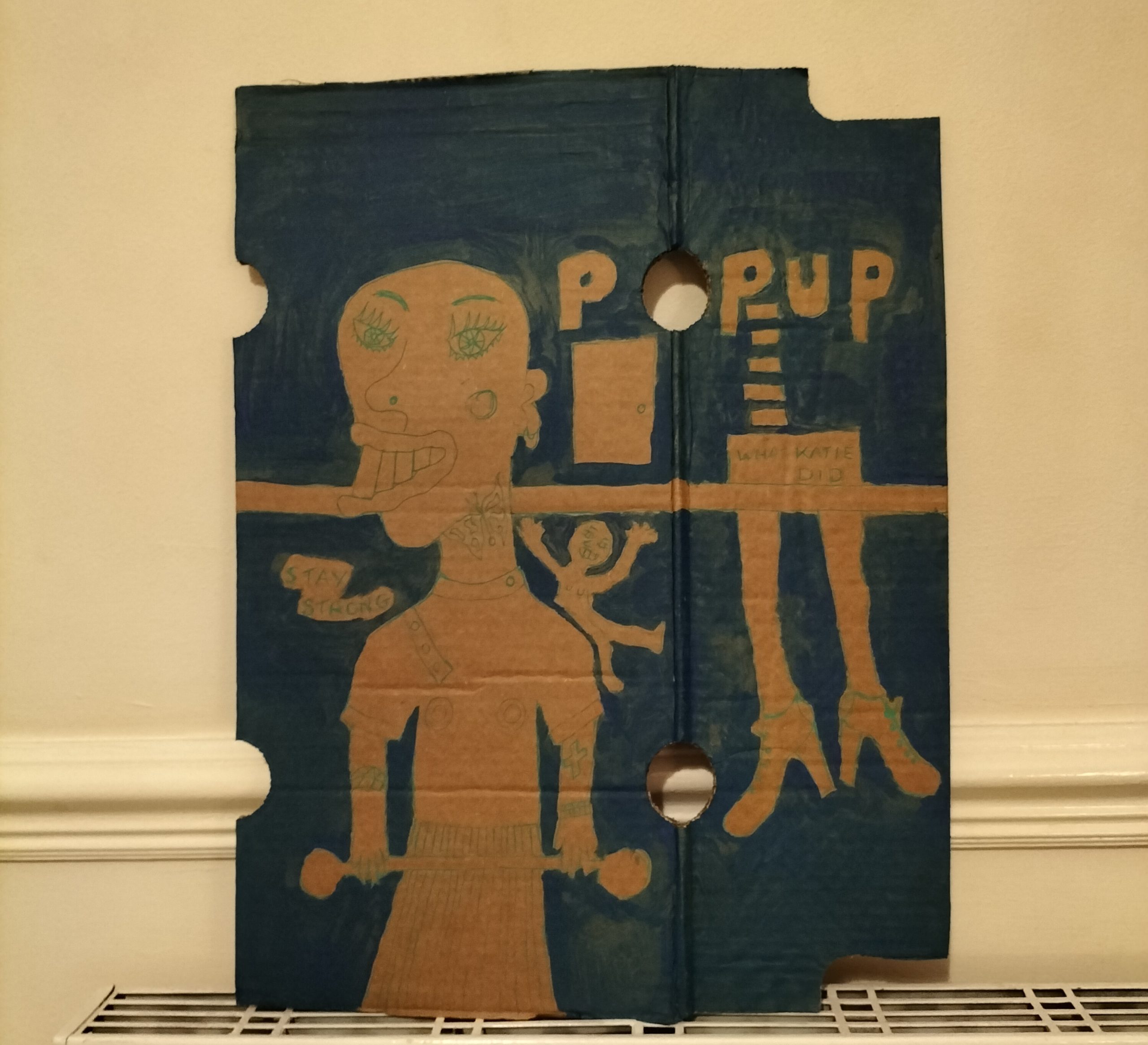 Cardboard art - What Katie did - blue background and figures drawn on the cardboard. On left a person lifting a weight with words Stay Strong and little figure on their shoulder. The words Pop Up around a hole in the cardboard. To the right waist and legs in shoes hanging from a bar going across the image and the words What Katie Did.