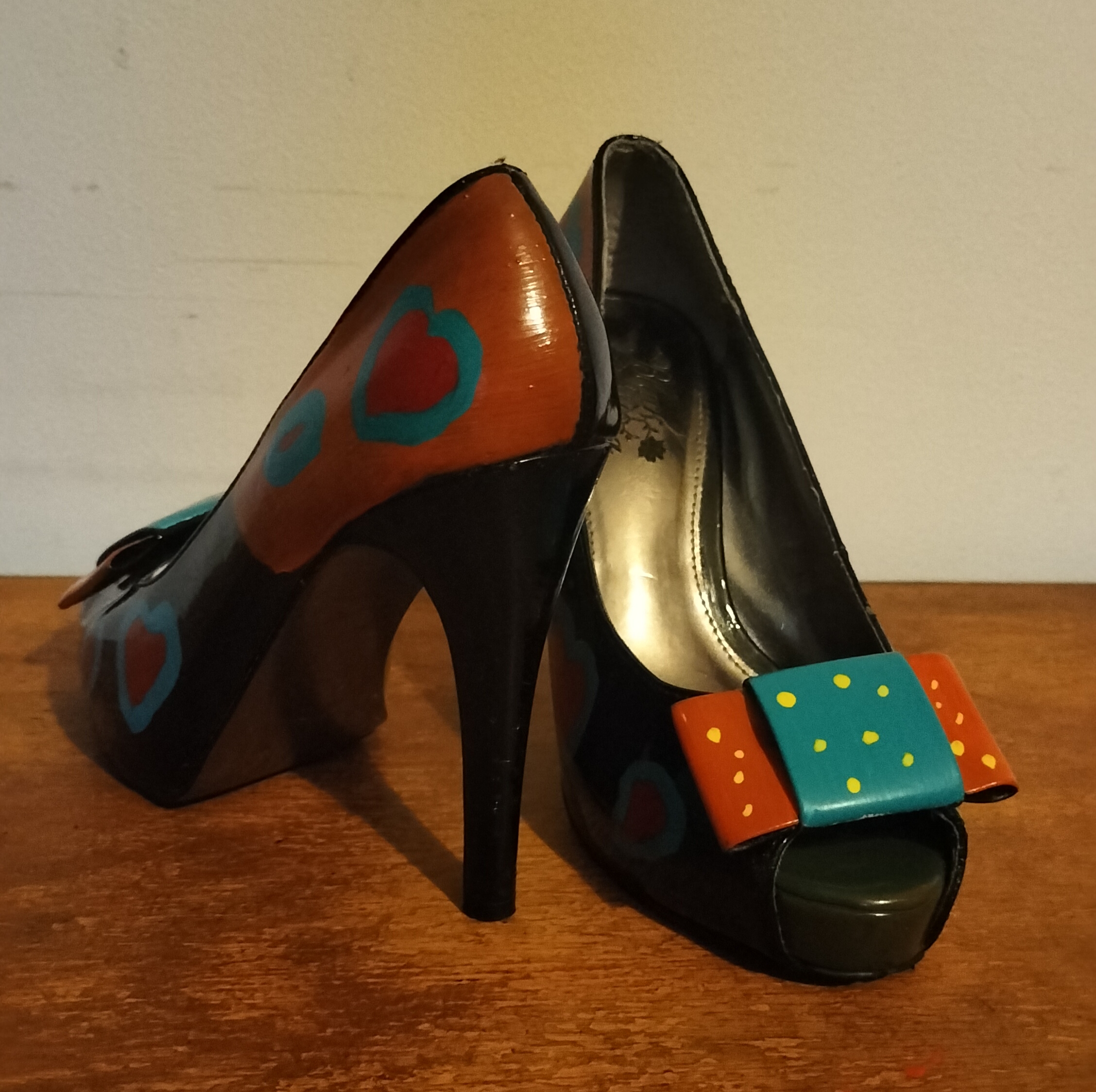 Size 3 or 4 peep toe shoes. Stiletto heels. Inside of shoes bottom half are painted yellow with red hearts surrounded in turquoise. Inside of upper part of the shoe is left plain apart from a couple of hearts in red with turquoise surround. Front of the shoes have bows. Hand painted too. Layered bows at the front and the lower layer is painted orangey/red with yellow spots and the top layer of the bow is turquoise with yellow spots. Outside of the shoe lower part is black shoe with hearts and the top half of the outside of the shoes are orange background with the red hearts surrounded in turquise. Small platform and open peep toe.