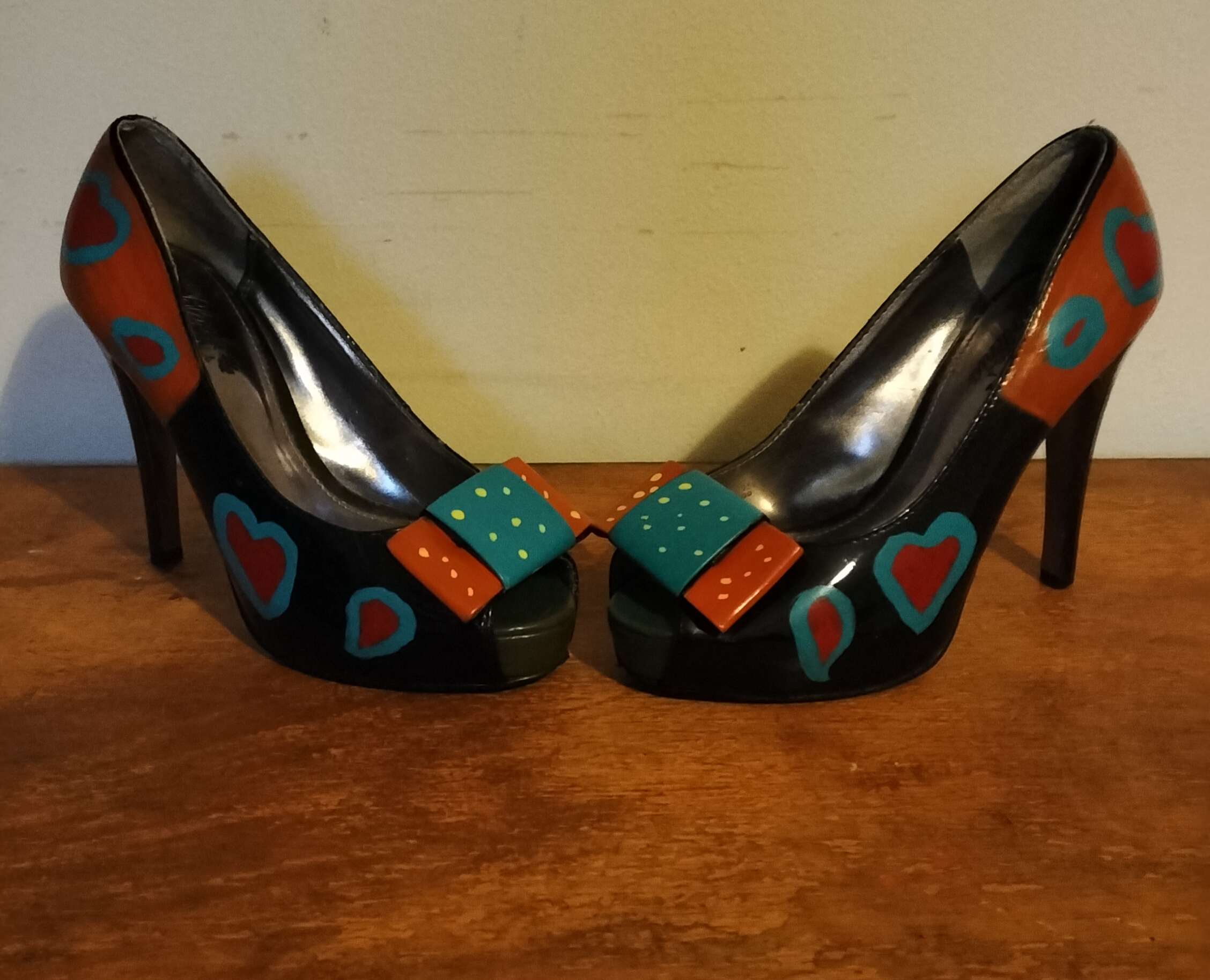 Size 3 or 4 peep toe shoes. Stiletto heels. Inside of shoes bottom half are painted yellow with red hearts surrounded in turquoise. Inside of upper part of the shoe is left plain apart from a couple of hearts in red with turquoise surround. Front of the shoes have bows. Hand painted too. Layered bows at the front and the lower layer is painted orangey/red with yellow spots and the top layer of the bow is turquoise with yellow spots. Outside of the shoe lower part is black shoe with hearts and the top half of the outside of the shoes are orange background with the red hearts surrounded in turquise. Small platform and open peep toe.