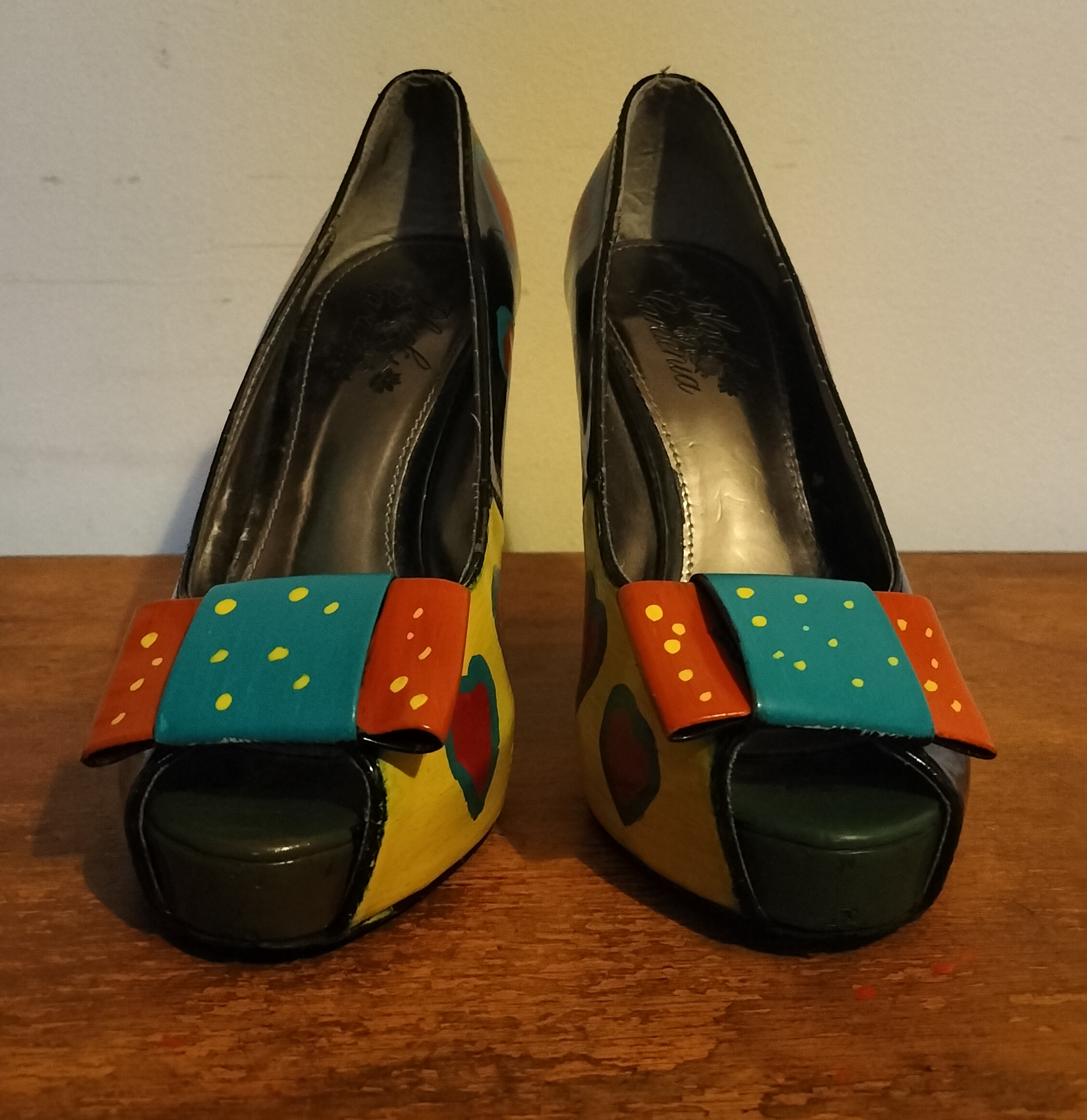 Size 3 or 4 peep toe shoes. Stiletto heels. Inside of shoes bottom half are painted yellow with red hearts surrounded in turquoise. Inside of upper part of the shoe is left plain apart from a couple of hearts in red with turquoise surround. Front of the shoes have bows. Hand painted too. Layered bows at the front and the lower layer is painted orangey/red with yellow spots and the top layer of the bow is turquoise with yellow spots. Outside of the shoe lower part is black shoe with hearts and the top half of the outside of the shoes are orange background with the red hearts surrounded in turquise. Small platform and open peep toe.