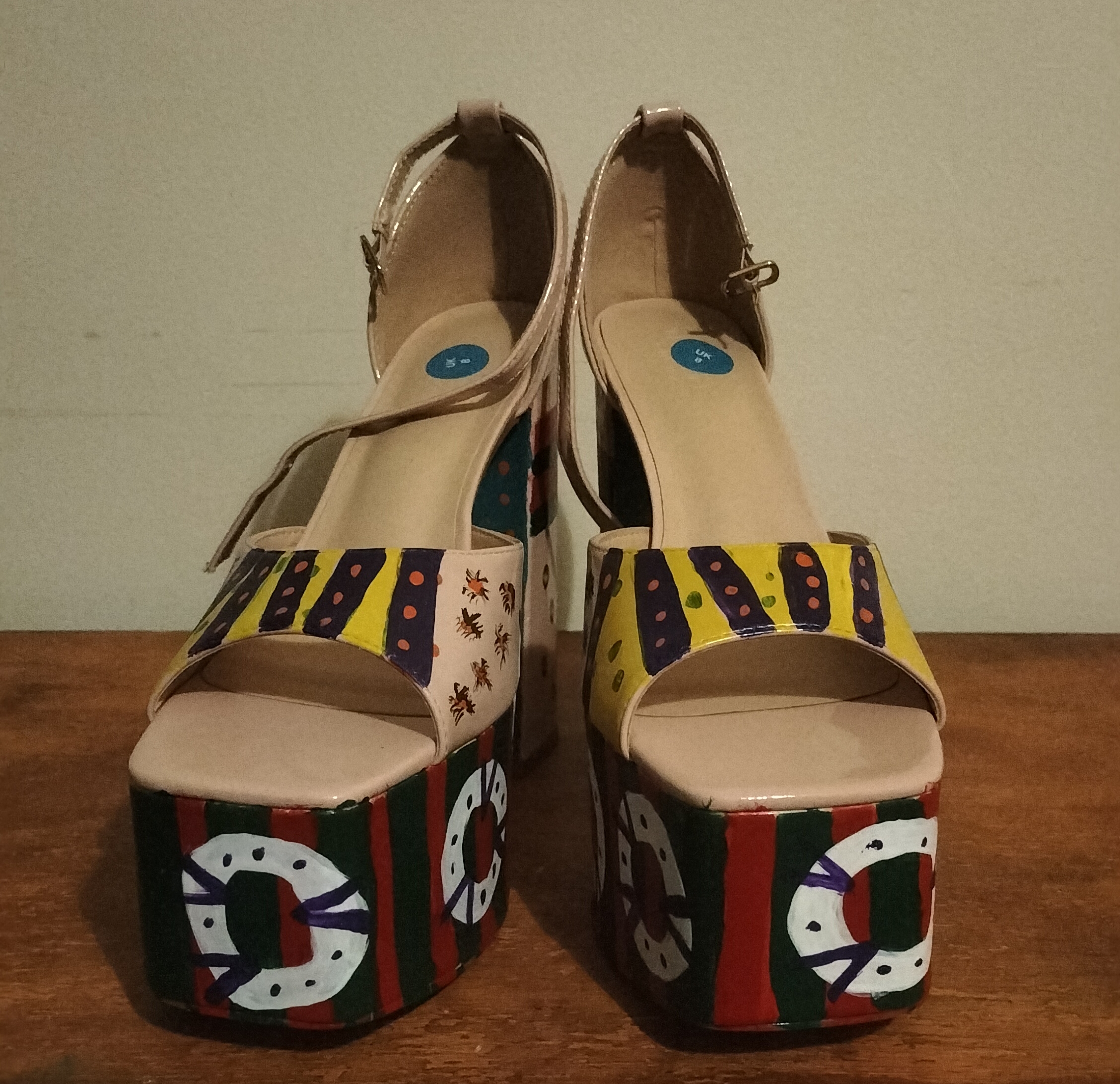 Size 8 preloved platform shoes. Beige uPVC handpainted. Proper funky and original. Green and red on the front platform (big) and then white circles with spots and lines. Turquoise hearts with yellow spots and black lines surrounding the hearts on the bottom of the heels and also on the back of the shoe behind your heel. Ankle straps. Open toe shoes. The section over the front of your foot is yellow and purple striped and there is more design on the side of the shoes. Original design.