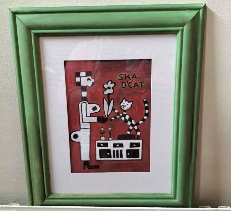 Ska d'cat original painting A4. Red background. Figure on the left holding a flower. Ska d'cat standing on a sideboard also holding the flower. Words read Ska d'cat. Black and white cat, signature black and white of ska/two tone music.