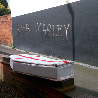 Coffin soft sculpture photographed on a wall with the words Bob Marley as graffiti on another wall behind it.