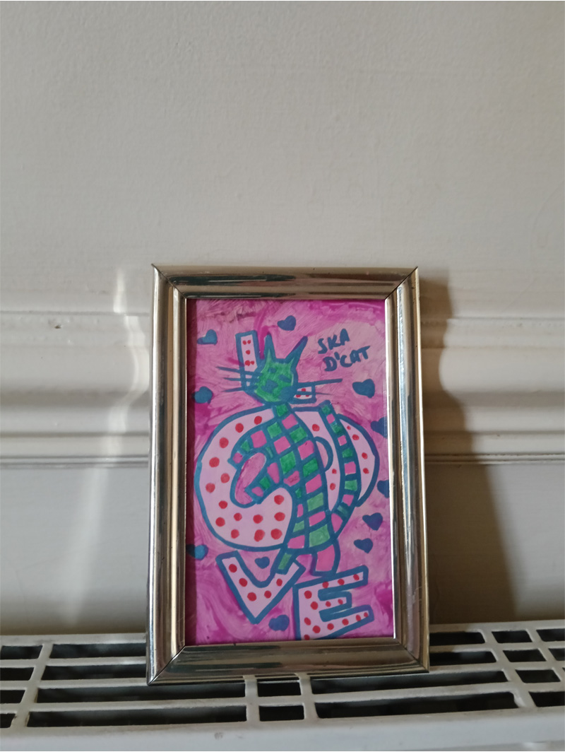 Little Ska d'cat framed. About 3 inches by 5 inches tall. Green and pink cat against lettering spelling Love. Standalone letters for the word "love" and the "l" is largely obscured by the cat. hearts on the image and the words "Ska d'cat". The letters for the word "love" are in pink with red dots.