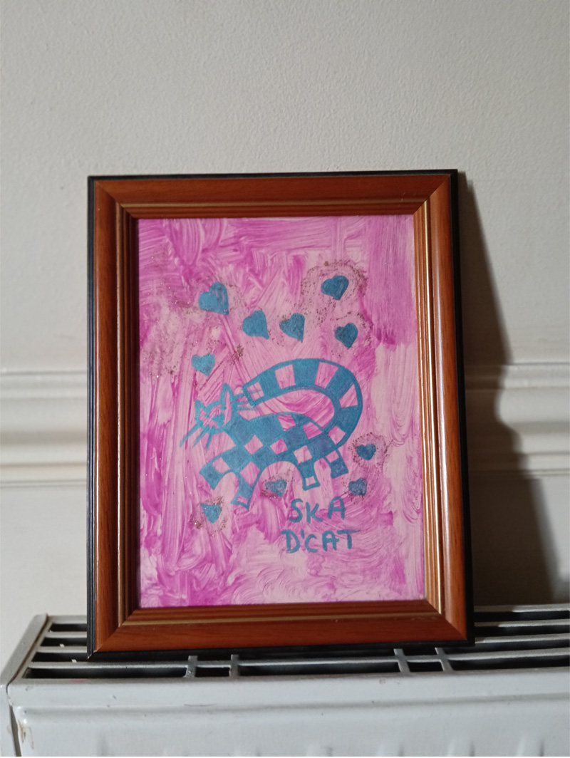 Ska dcat about 4 inches by 6 inches framed. Pink paint background, uneven surface. Grey cat which has squares on it. Alternative squares are filled in with colour like the two tone for ska music. Grey hearts on the image and the words "Ska d'cat". A play on words for lovers of ska music and cats.