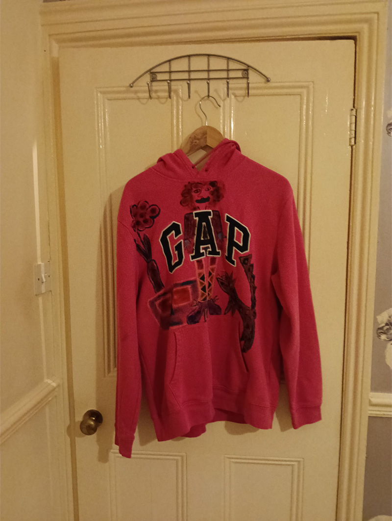 Gap hoodie size 16. Pink. Hand painted on the front. Woman figure around the A of the word Gap. Shows gap at top of legs which is referencing a woman's weight and space between her thighs. Sideboard to the left of the legs and shoes. Knee length socks. Dog to the right of the legs. Plant with leaves and flower on top of the sideboard. Vintage hoodie. Painted front and back. Back of hoodie has a star ont he hood and a figure on the back of the hoodie which is abstract, bald headed and the words Reach for the Stars appear in a black line surround.