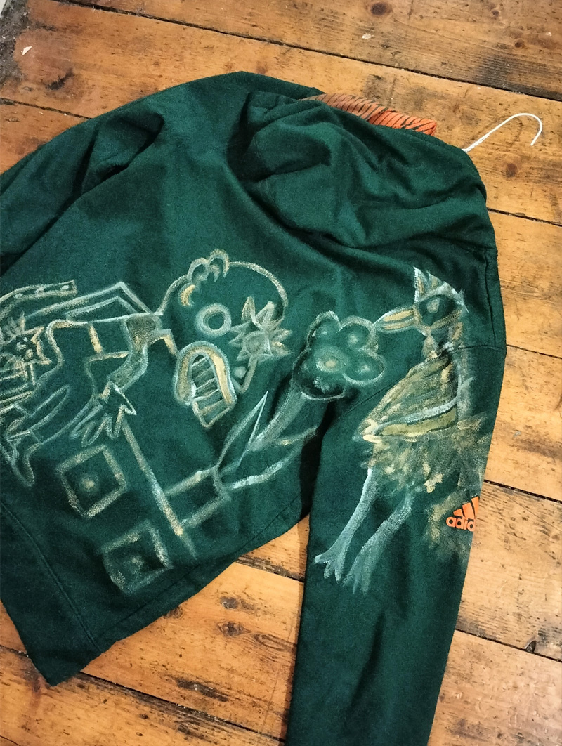 Dark green Adidas hoodie, small. Hand painted front and back and sleeves. Front has some kind of face-like image. The back has a seated figure, a cat and sideboard with a plant on it. On one of the shoulders/arms is a bird and this image also comes onto the back of the shoulder. On the other arm is an abstract dog. All images are outlined in gold paint and part of the art work is filled in with gold paint.
