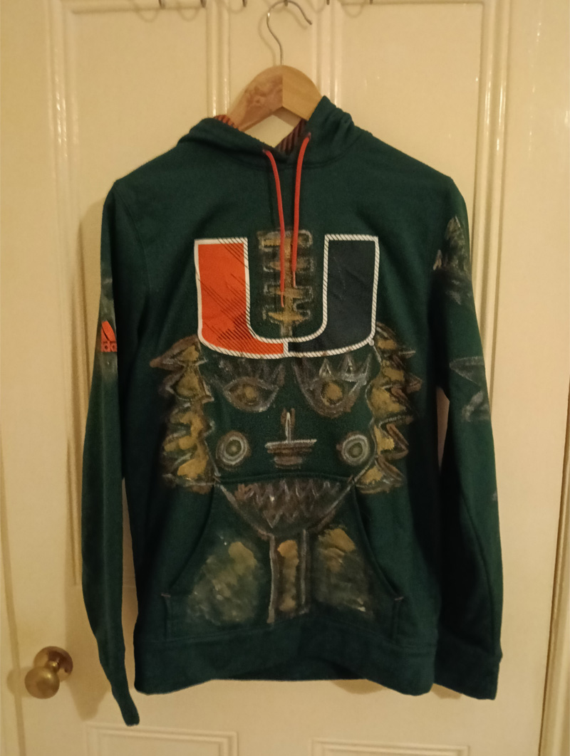 Dark green Adidas hoodie, small. Hand painted front and back and sleeves. Front has some kind of face-like image. The back has a seated figure, a cat and sideboard with a plant on it. On one of the shoulders/arms is a bird and this image also comes onto the back of the shoulder. On the other arm is an abstract dog. All images are outlined in gold paint and part of the art work is filled in with gold paint.