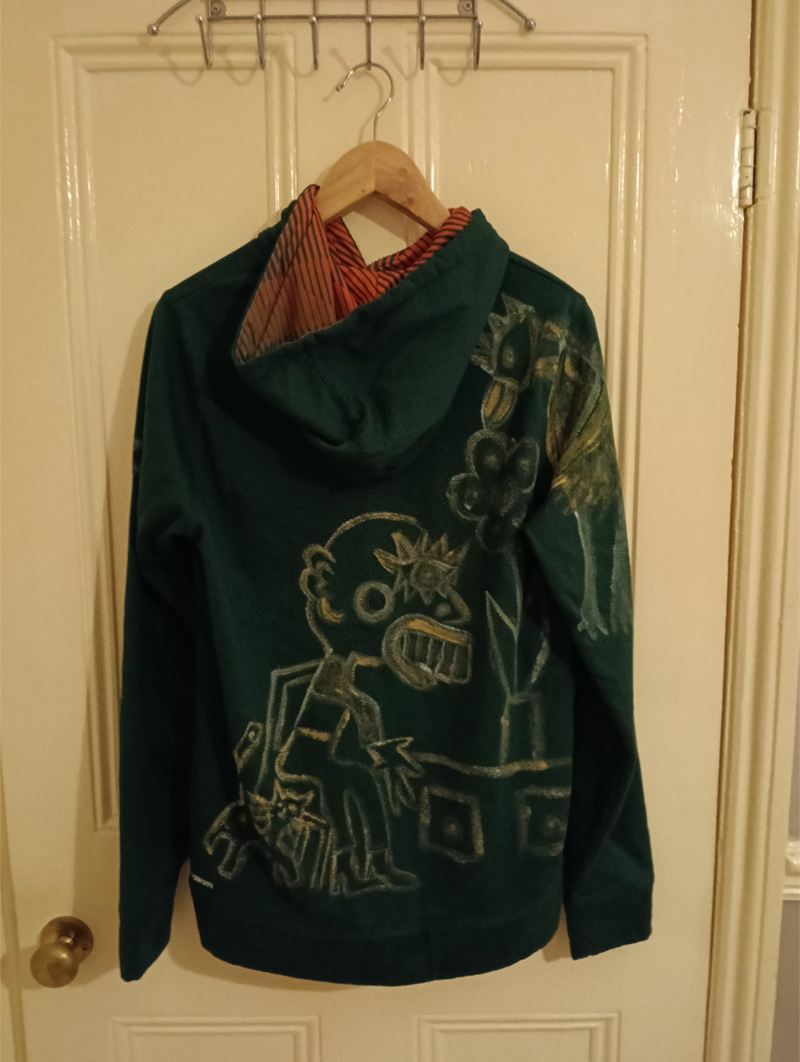 Dark green Adidas hoodie, small. Hand painted front and back and sleeves. Front has some kind of face-like image. The back has a seated figure, a cat and sideboard with a plant on it. On one of the shoulders/arms is a bird and this image also comes onto the back of the shoulder. On the other arm is an abstract dog. All images are outlined in gold paint and part of the art work is filled in with gold paint.