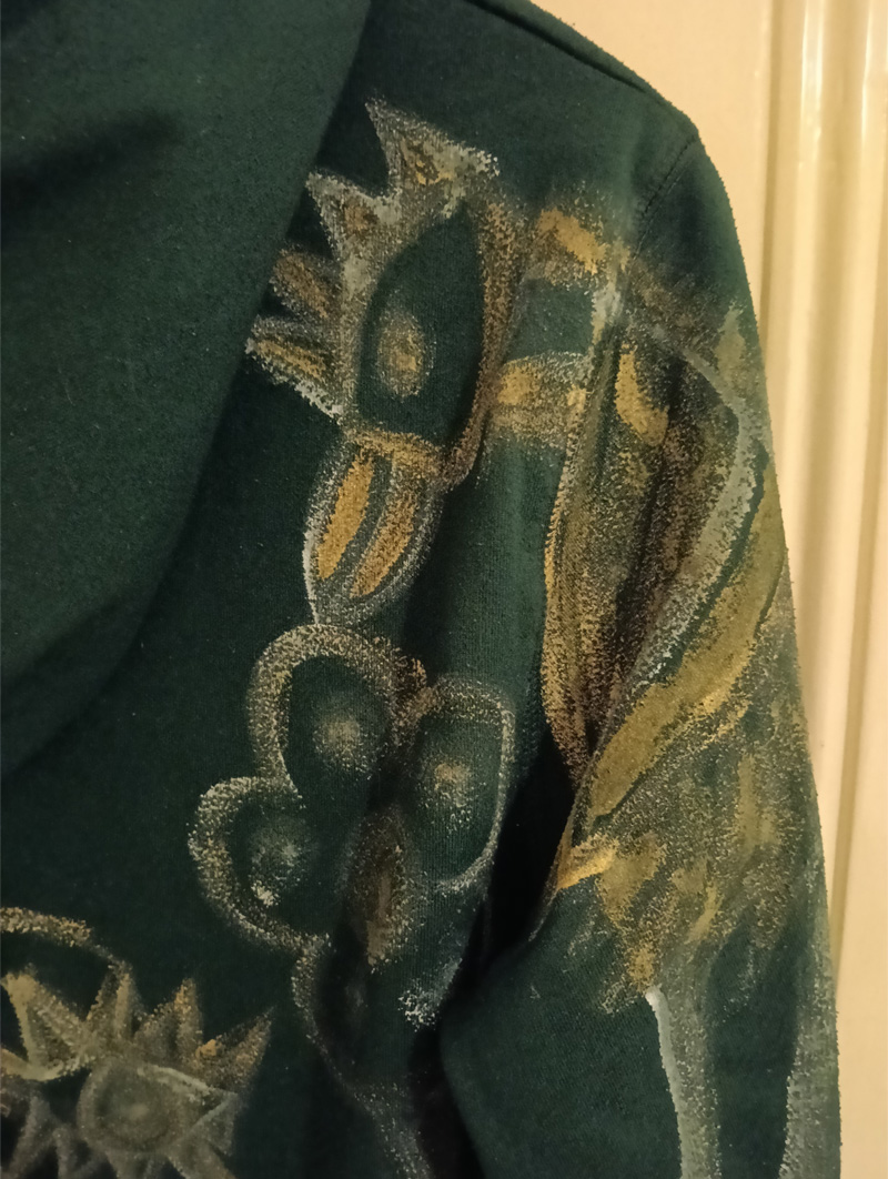 Dark green Adidas hoodie, small. Hand painted front and back and sleeves. Front has some kind of face-like image. The back has a seated figure, a cat and sideboard with a plant on it. On one of the shoulders/arms is a bird and this image also comes onto the back of the shoulder. On the other arm is an abstract dog. All images are outlined in gold paint and part of the art work is filled in with gold paint.
