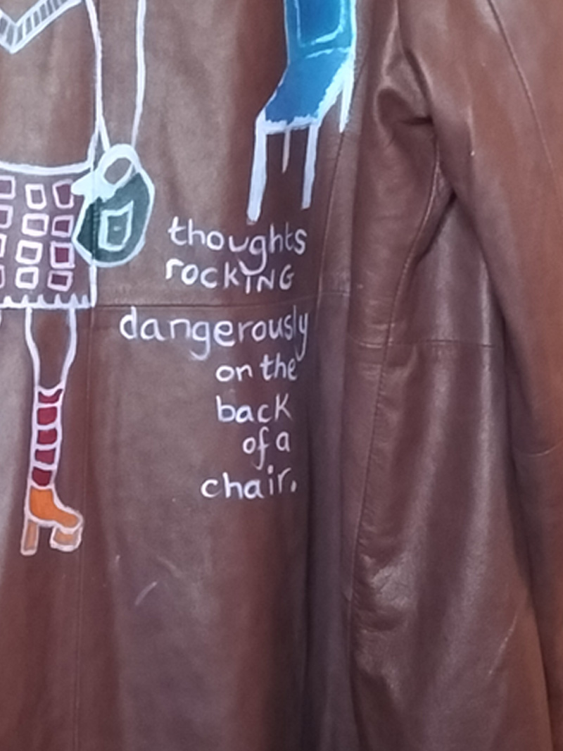 Back of jacket is hand painted. Figure on the left is wearing a skirt and top. Bald headed and carrying a handbag. Knee high socks and orange platform shoes. Smaller figure balancing on the main figure's shoulder and the top of a high backed chair. Similar shoes and trousers and top. Also bald headed. The words "Oxford Bags" across the trousers and between the legs below reads "+ easy shags". Blue chair. Words underneath the chair read "thoughts rocking dangerously on the back of a chair." Only painted on the back of the jacket. Buttoned up front. A slanted pocket to the left and right of the jacket. Arma leather jacket size 16. Brown.
