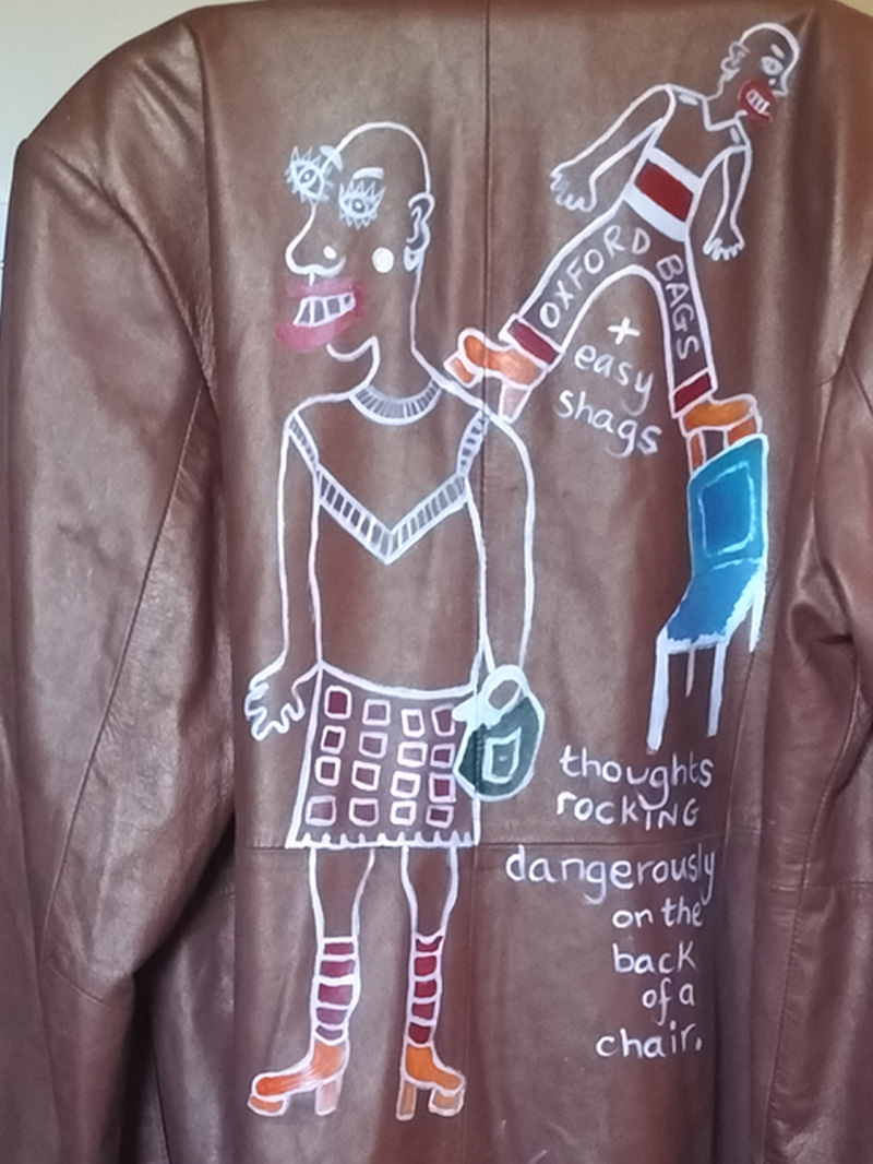 Back of jacket is hand painted. Figure on the left is wearing a skirt and top. Bald headed and carrying a handbag. Knee high socks and orange platform shoes. Smaller figure balancing on the main figure's shoulder and the top of a high backed chair. Similar shoes and trousers and top. Also bald headed. The words "Oxford Bags" across the trousers and between the legs below reads "+ easy shags". Blue chair. Words underneath the chair read "thoughts rocking dangerously on the back of a chair." Only painted on the back of the jacket. Buttoned up front. A slanted pocket to the left and right of the jacket. Arma leather jacket size 16. Brown.