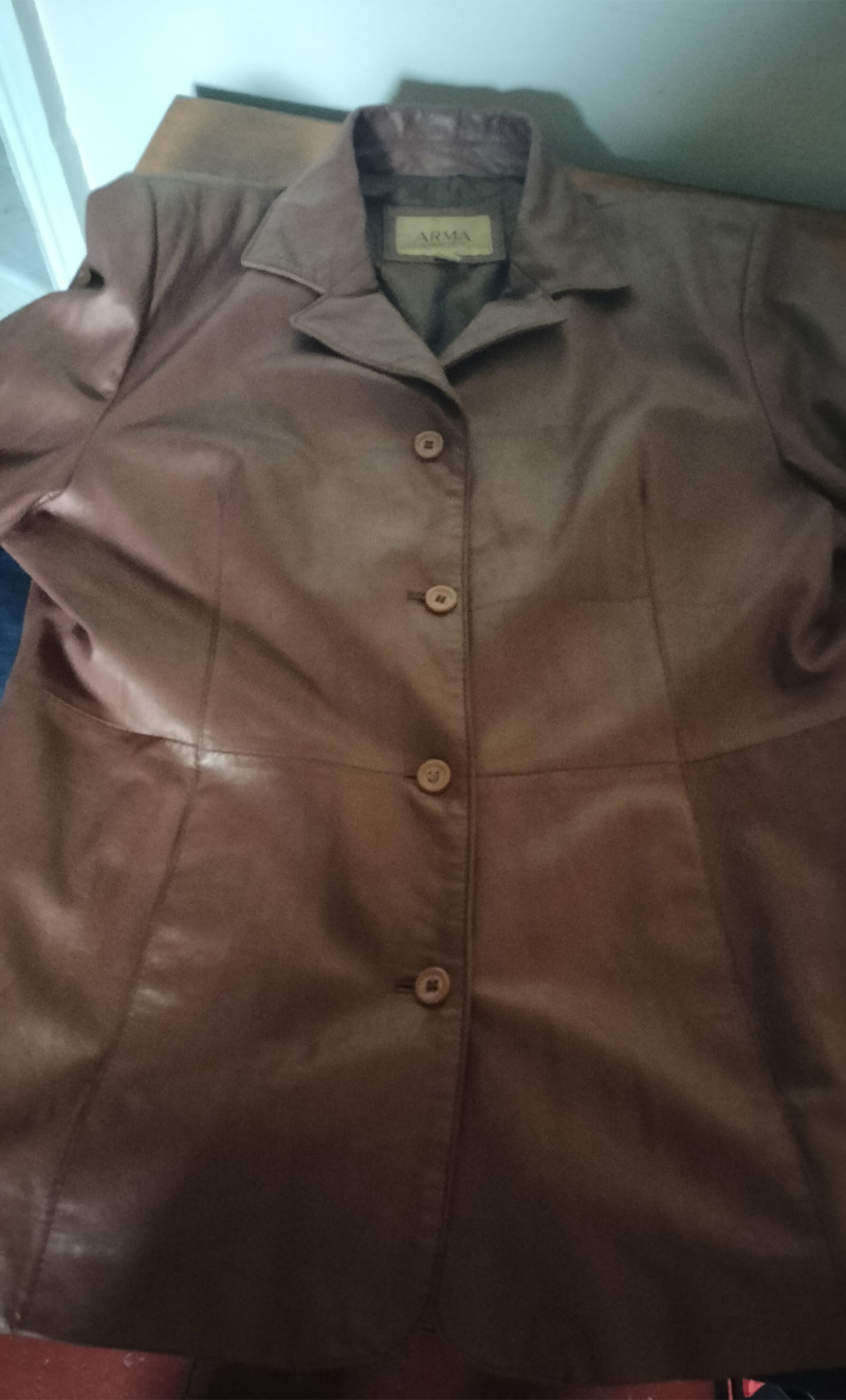 Back of jacket is hand painted. Figure on the left is wearing a skirt and top. Bald headed and carrying a handbag. Knee high socks and orange platform shoes. Smaller figure balancing on the main figure's shoulder and the top of a high backed chair. Similar shoes and trousers and top. Also bald headed. The words "Oxford Bags" across the trousers and between the legs below reads "+ easy shags". Blue chair. Words underneath the chair read "thoughts rocking dangerously on the back of a chair." Only painted on the back of the jacket. Buttoned up front. A slanted pocket to the left and right of the jacket. Arma leather jacket size 16. Brown.