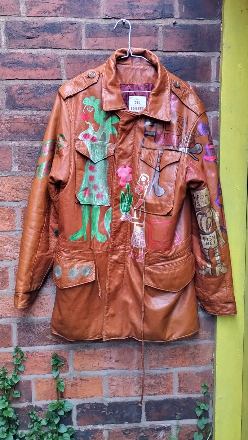 Liam's Old Coat - hand painted vintage parka jacket with hood (brown). Full image of the front of the jacket. Hand painted with different images. Green woman image on the right side of the front of the jacket. To the left mid height is a character holding a flower with leaves. Another abstract figure around the top left pocket. Circles on the flap over the right bottom pocket. Vintage Rome Peabody Massachusetts USA Size 42 brown leather hooded parka jacket handpainted one off.