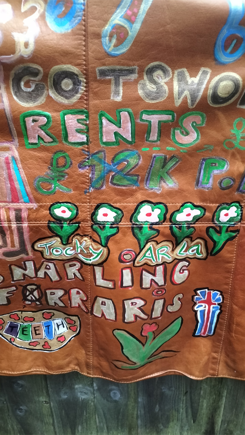 Liam's Old Coat - hand painted vintage parka jacket with hood (brown). Close up of words on the back of jacket. Tocky Ar La (purposely misspelt), Cotswolds Rents £12k pm (crossed out) and replaced by £19k pm. Snarling Farraris (misspelt) below it Teeth illustration (teeth with the lettering spelling "teeth" on the teeth) pointing one of the "e" letters to the Farrari word. This is the bottom of the back of the jacket. There is more illustration at the top of the jacket. Vintage Rome Peabody Massachusetts USA Size 42 brown leather hooded parka jacket handpainted one off.