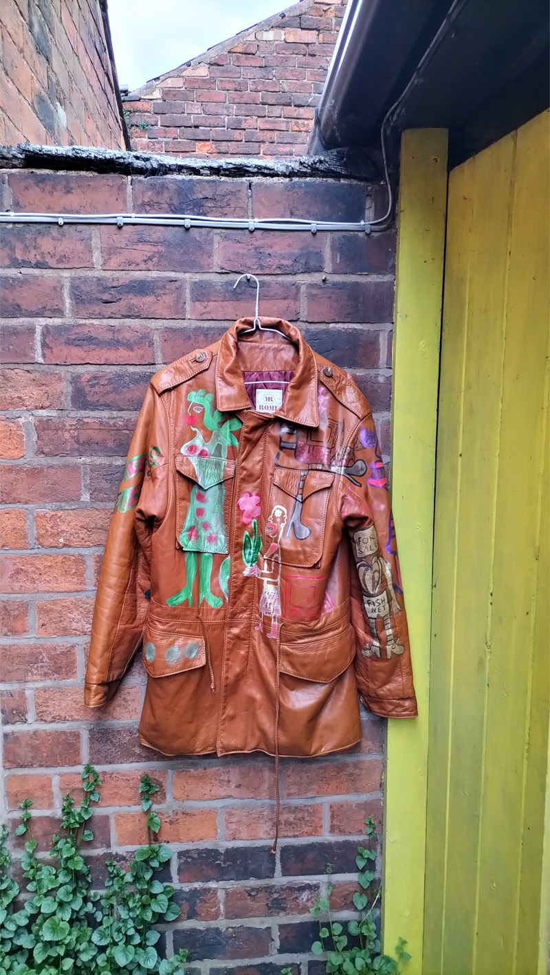 Liam's Old Coat - hand painted vintage parka jacket with hood (brown). Full image of the front of the jacket. Hand painted with different images. Green woman image on the right side of the front of the jacket. To the left mid height is a character holding a flower with leaves. Another abstract figure around the top left pocket. Circles on the flap over the right bottom pocket. Vintage Rome Peabody Massachusetts USA Size 42 brown leather hooded parka jacket handpainted one off.