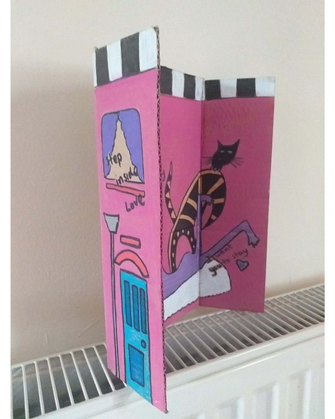 One for the lovers of Cilla Black piece uses some of the words of her song. Step inside love - and stay, I want you to stay cardboard art. About A4 landscape in size folded into three panels. When closed the front of the piece is like the front of a house. When opened it is a image of a purple woman on a rug with a cat and a plant. Front reads Step inside love. Inside reads and stay and lower down the image reads I want you to stay. The back of the image when opened out has painted parts on the left and right of the card and the centre piece is plain cardboard with a sticker on it. Step inside love on the front of the piece. Inside reads … and stay … I want you to stay.