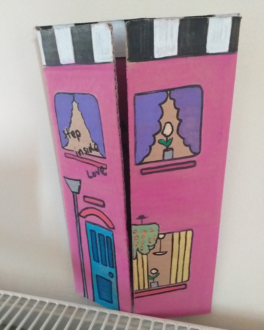 One for the lovers of Cilla Black piece uses some of the words of her song. Step inside love - and stay, I want you to stay cardboard art. About A4 landscape in size folded into three panels. When closed the front of the piece is like the front of a house. When opened it is a image of a purple woman on a rug with a cat and a plant. Front reads Step inside love. Inside reads and stay and lower down the image reads I want you to stay. The back of the image when opened out has painted parts on the left and right of the card and the centre piece is plain cardboard with a sticker on it. Step inside love on the front of the piece. Inside reads … and stay … I want you to stay.