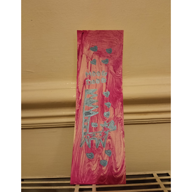 Original hand painted Moog Doog bookmark. Grey outline for the dog. Grey hearts above and below the dog and the words Moog Doog above the dog. Pink background. Keyboard tail straight on the left and zig zag on the right. Grey outline for the dog body and head. Spots on the body representing the dials on the sythesizer. Design is based on the Robert Moog synthesizer known as the Moog Synthesizer.