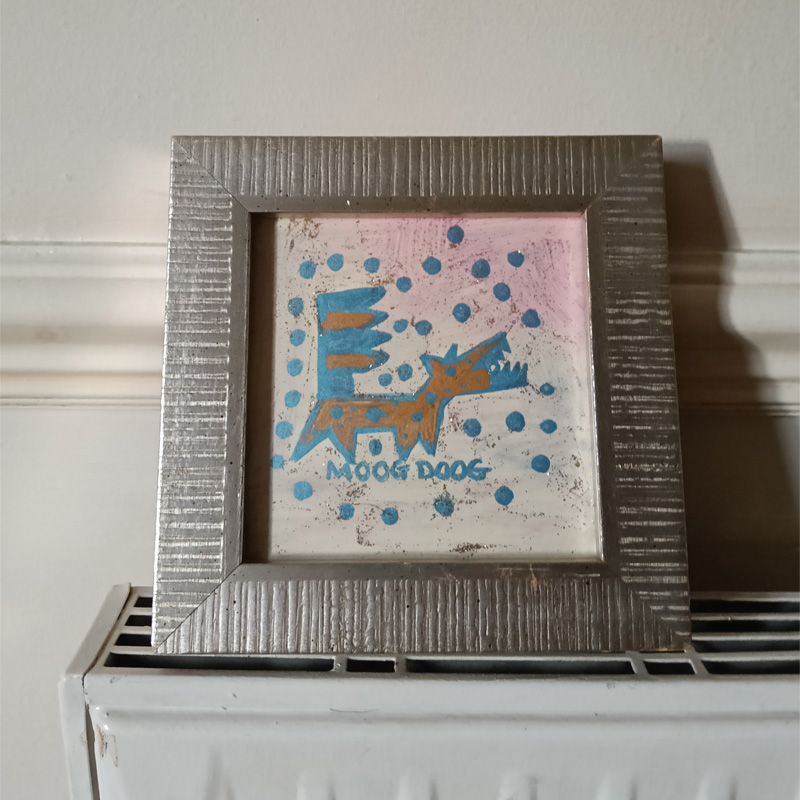 Small framed original. Silver frame. Moog Doog hand painted piece. The dog is cartoon like based on the Moog synthesizer. A keyboard for a tail, straight on the left side and zig zag on the right. The body of the dog has circles and dots on it representing the dials on the synthesizer. Blue and brown colours. Blue dots around the dog and the words Moog Doog below the dog.