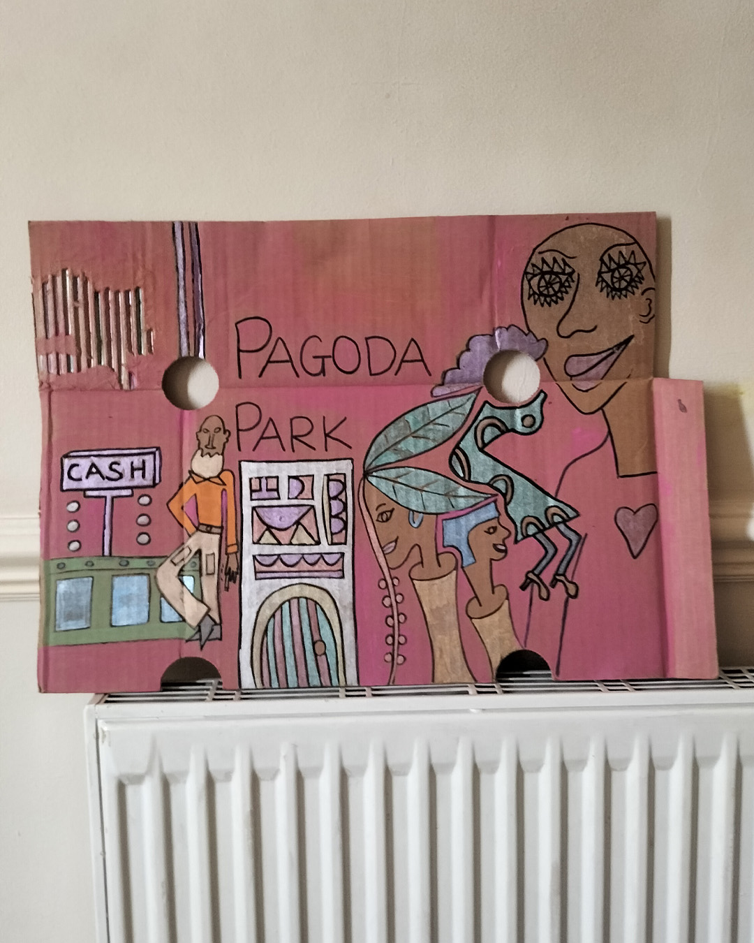 Night club scene on A3 cardboard. Man at cash counter and couple of heads coming out of what could be tree trunks. Two holes in the cardboard in the top third of the cardboard. One hone on the right is used as an empty face of a woman. Large figure at the top right of the cardboard. Head and shoulders and heart shape on the top of the person. Image that could be a fancy door with glass panels. Bit of the cardboard towards the top left has its top surface torn off. With the two heads that are coming out of tree trunks, one has leaves as hair. The words Pagoda Park on the cardboard.