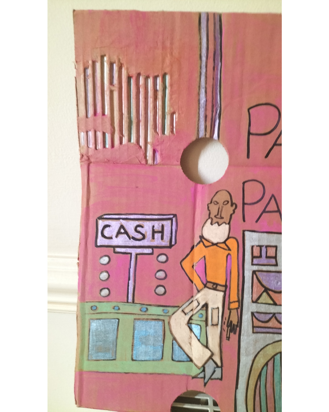 Night club scene on A3 cardboard. Man at cash counter and couple of heads coming out of what could be tree trunks. Two holes in the cardboard in the top third of the cardboard. One hone on the right is used as an empty face of a woman. Large figure at the top right of the cardboard. Head and shoulders and heart shape on the top of the person. Image that could be a fancy door with glass panels. Bit of the cardboard towards the top left has its top surface torn off. With the two heads that are coming out of tree trunks, one has leaves as hair. The words Pagoda Park on the cardboard.