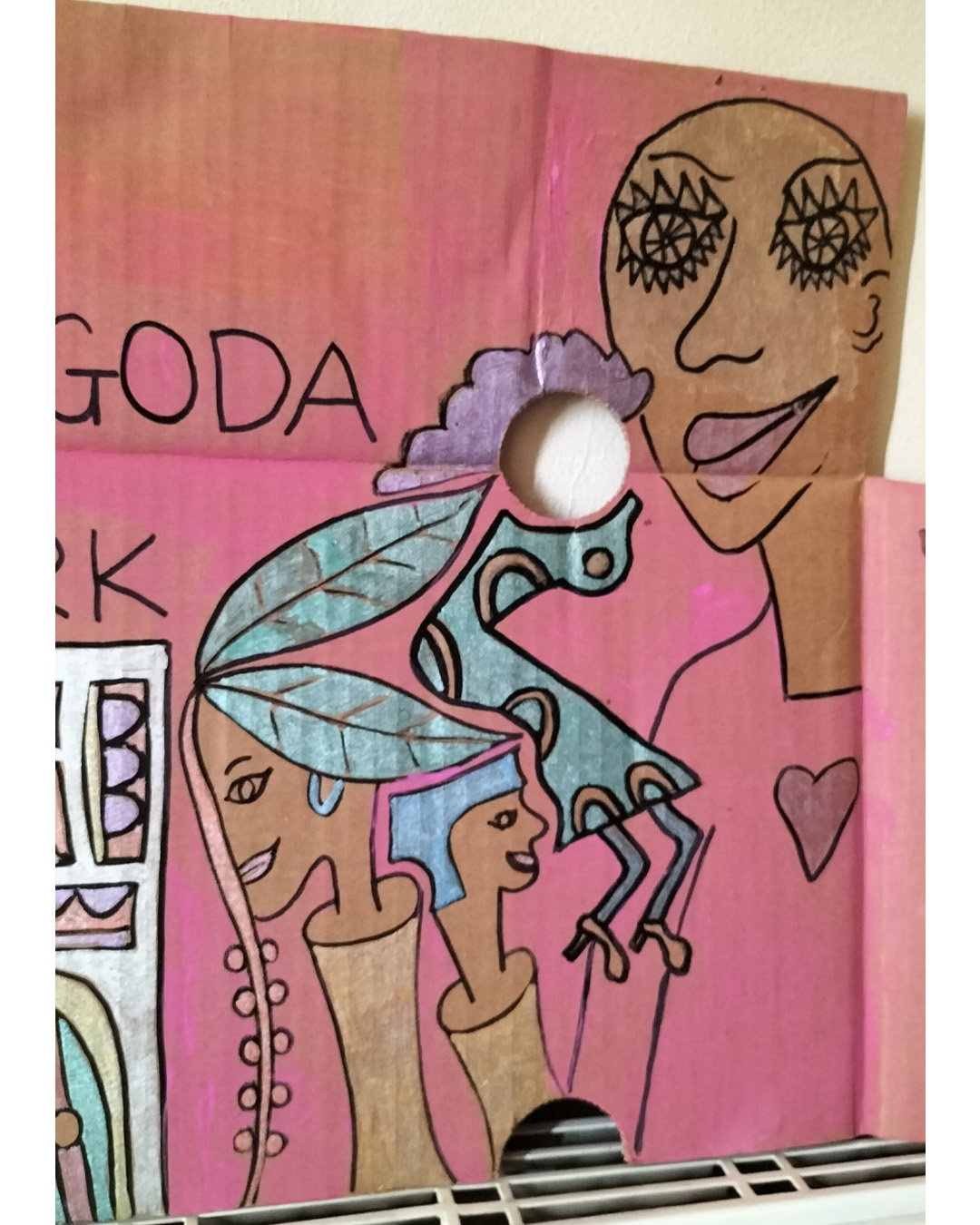 Night club scene on A3 cardboard. Man at cash counter and couple of heads coming out of what could be tree trunks. Two holes in the cardboard in the top third of the cardboard. One hone on the right is used as an empty face of a woman. Large figure at the top right of the cardboard. Head and shoulders and heart shape on the top of the person. Image that could be a fancy door with glass panels. Bit of the cardboard towards the top left has its top surface torn off. With the two heads that are coming out of tree trunks, one has leaves as hair. The words Pagoda Park on the cardboard.